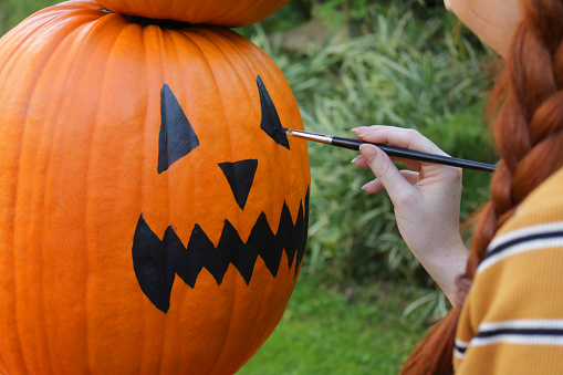 Close-up image of red-haired, young woman using paintbrush to paint faces on pumpkins, stack of orange gourds, Halloween face Jack O'Lantern outdoors in garden