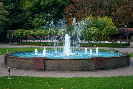 Big city beautiful working fountain in a public place in the bright autumn sun. Trees and bushes in the parking