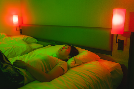A young woman is lit by red and green neon colored lights and resting in bed in bedroom at home.