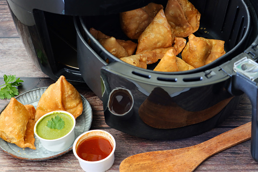 Close-up image of of cooking pan of fried samosas, stuffed with spiced potato, peas and meat, removed from air fryer, besides ramekins of mint coriander dip and mango chutney, healthier alternative to frying pan cooking, elevated view, focus on foreground