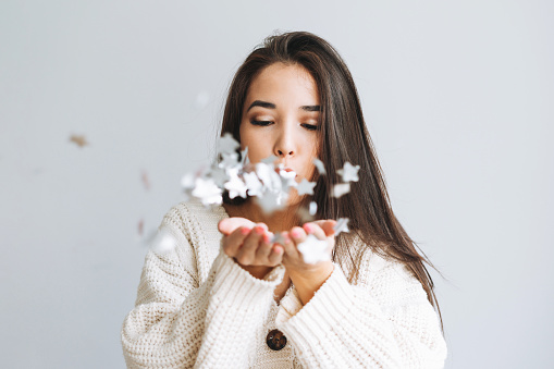 Young asian woman in white knitted cardigan blows on snowflakes in hands on grey background, winter christmas time