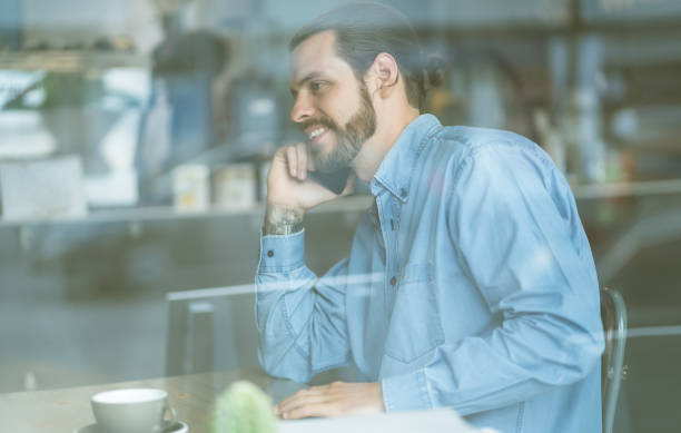 istock Stylish male freelancer talking on the mobile phone and smiling while sitting at table behind glass in cafe.Cozy office workplace. 1346855280