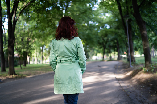 One redhead woman in green coat enjoying relaxing walk in city park on sunny day, back view