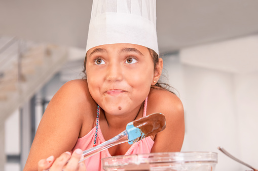 Close up view of Latina girl in a chef's hat grimaces as she prepares a chocolate cake at home