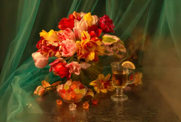 Photo of Still life with a bouquet of colorful tulips in a transparent vase on a green background. Cup with tea and marmelade
