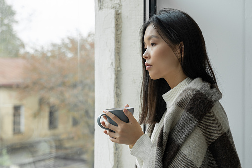 Young sad Asian woman drinks coffee or tea and looks out the window covered with a blanket.