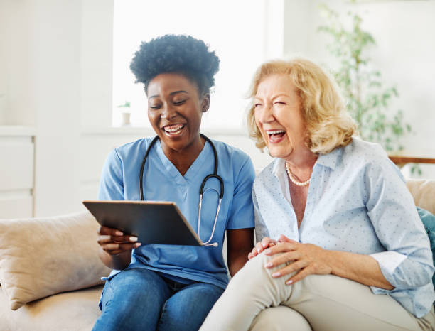 nurse doctor senior care tablet computer technology showing caregiver help assistence retirement home nursing elderly woman african american black happy laughing Doctor or nurse caregiver showing a tablet screen to  senior woman and laughing at home or nursing home community outreach stock pictures, royalty-free photos & images