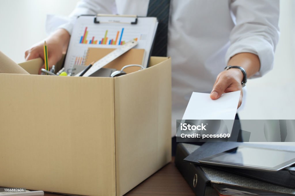 Resignation. Businessman has a brown cardboard box next to his body and sends a letter of resignation to the employer boss. resignation letter staff reduction resigning leaving the office Quitting a Job Stock Photo