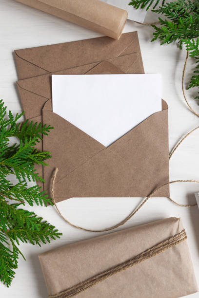 istock Festive background from natural material for Christmas and New Year. Opened craft envelope with a blank sheet of paper and wrapped gifts tied with a rope on a white wooden table. 1346883610