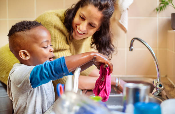 Smiling little boy and his mom washing dishes together in their kitchen stock photo