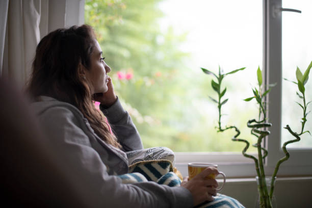 mujer deprimida sentada junto a la ventana - tristeza fotografías e imágenes de stock