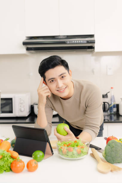 istock Young asian man holding apple fruit and using tablet while standing in kitchen room 1346889124