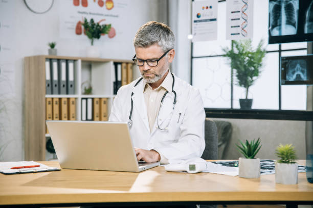 istock Grey haired doctor sitting at desk and typing on laptop 1346890402