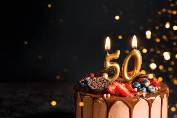 Chocolate birthday cake with berries, cookies and number fifty golden candles on black background, copy space.