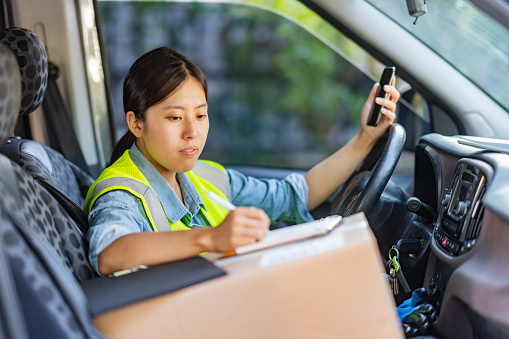 A female delivery person is sitting in a van with parcels next to her and using a smart phone to check and update orders.