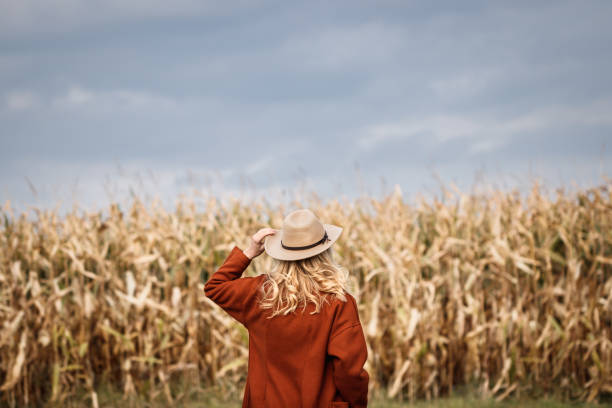 istock Stylish woman wearing red coat and hat is standing next corn field 1346905697