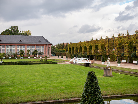 Schwetzingen, Germany - October 13, 2021: Schwetzingen Castle Park. Orangery, built by Karl Theodor. The walls are decorated with facade paintings and simulate natural stone. In the foreground a green meadow with statues framed by a small creek.