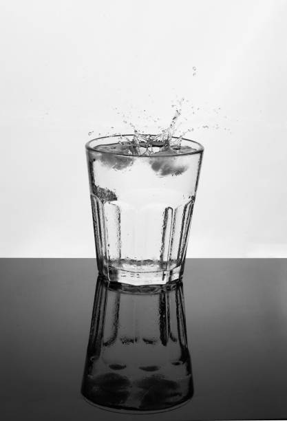 Glass of water on a reflective surface splashing drops, white background. Drinks and hydration concept