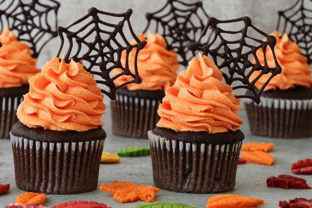 Close-up image of batch homemade, Halloween chocolate cupcakes in paper cake cases with orange butter icing piped swirls topped with chocolate spider's webs, fondant autumnal coloured leaves on marble effect background Stock photo showing a close-up view of freshly baked, homemade, chocolate cupcakes in paper cake cases. The cup cakes have been decorated with swirls of orange coloured piped icing and chocolate cobwebs. halloween cupcake stock pictures, royalty-free photos & images