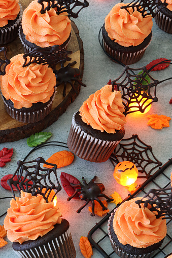 Stock photo showing a close-up view of a batch of freshly baked, homemade, chocolate cupcakes in paper cake cases on wood and metal cake stand. The cup cakes have been decorated with swirls of orange coloured piped icing and chocolate cobwebs.