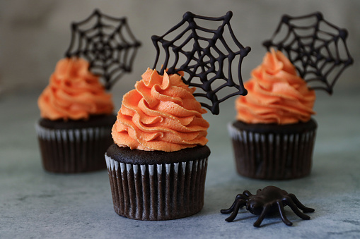 Stock photo showing a close-up view of freshly baked, homemade, chocolate cupcakes in paper cake cases. The cup cakes have been decorated with swirls of orange coloured piped icing and chocolate cobwebs.
