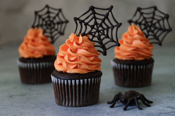 Close-up image of three homemade, Halloween chocolate cupcakes in paper cake cases with orange butter icing piped swirls topped with chocolate spider's webs, plastic spider on marble effect background Stock photo showing a close-up view of freshly baked, homemade, chocolate cupcakes in paper cake cases. The cup cakes have been decorated with swirls of orange coloured piped icing and chocolate cobwebs. halloween cupcake stock pictures, royalty-free photos & images