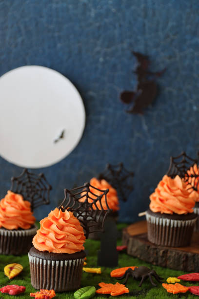 istock Image of Halloween graveyard scene displaying batch homemade, chocolate cupcakes in paper cake cases with orange butter icing piped swirls topped with chocolate spider's webs, fondant autumnal coloured leaves, flying witch and full moon blue background 1346918624