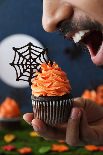 Stock photo showing a close-up view of freshly baked, homemade, chocolate cupcakes in paper cake cases decorated with swirls of orange coloured piped icing and chocolate cobwebs, displayed in a Halloween night scene of cemetery with full moon, witch flying a broomstick and gravestones on artificial grass surface.