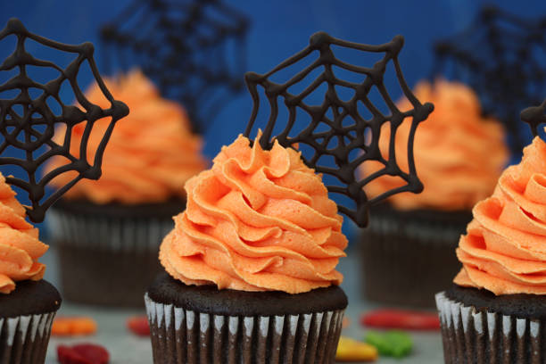istock Full frame image of batch homemade, Halloween chocolate cupcakes in paper cake cases with orange butter icing piped swirls topped with chocolate spider's webs, fondant autumnal coloured leaves on marble effect surface, blue background 1346918724
