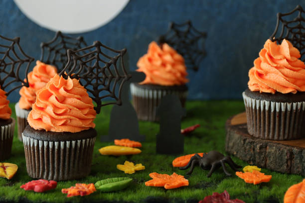 istock Close-up image of Halloween graveyard scene displaying batch homemade, chocolate cupcakes in paper cake cases with orange butter icing piped swirls topped with chocolate spider's webs, fondant autumnal coloured leaves, full moon blue background 1346918746