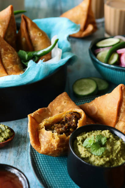 istock Image of plate of fried samosas one with missing bite, bowl lined with turquoise muslin, mint coriander dip and mango chutney ramekins, bowl salad vegetables, glasses of chai tea, blue wood grain surface, blue marble effect background, focus on foreground 1346923368