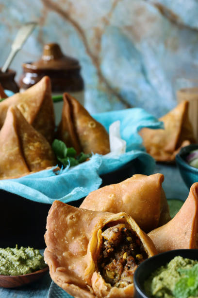 istock Image of plate of fried samosas one with missing bite, bowl lined with turquoise muslin, mint coriander dip and mango chutney ramekins, bowl salad vegetables, glasses of chai tea, blue wood grain surface, blue marble effect background, focus on foreground 1346923492
