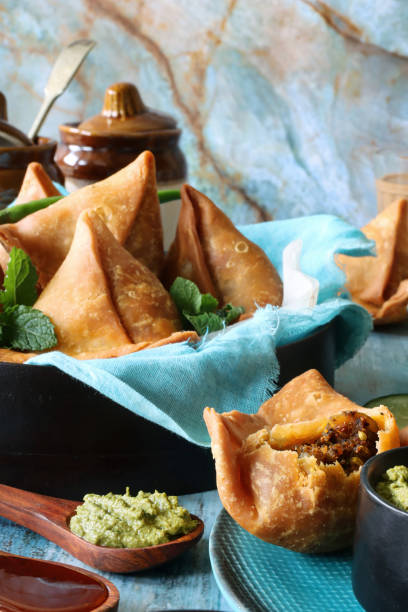 istock Image of fried samosa one with missing bite on plate, bowl lined with turquoise muslin, mint coriander dip ramekin, ceramic pickle lidded pots, glass of chai tea, wooden spoon, blue wood grain surface, blue marble effect background, focus on foreground 1346923553