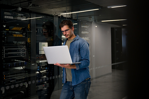 Latin American IT support technician fixing a problem in the server room using his laptop computer