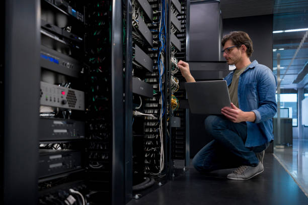 A man in glasses, wearing a blue shirt and casual pants, crouches beside a bank of servers, using a laptop to work on network connections in a modern data center.