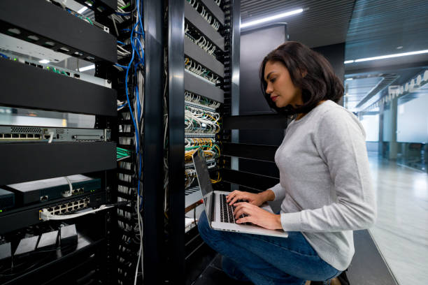 A woman with shoulder-length dark hair is crouched beside a server rack, working on a laptop surrounded by various networking equipment and colorful cables in a modern office environment.