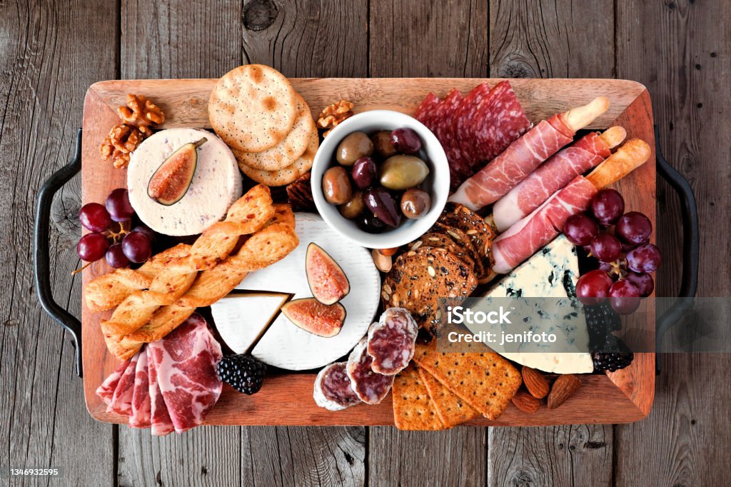 Charcuterie tray of a variety of meats, cheeses and appetizers over dark wood Charcuterie tray of a variety of meats, cheeses and appetizers. Top view on a dark wood background. Cheese Board Stock Photo