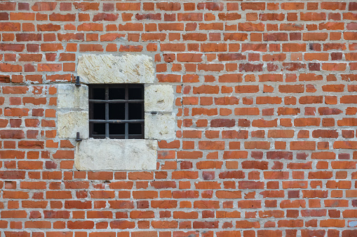 The old red brick fortress wall. There is a square window with a stone frame and iron bars. Background. Texture.