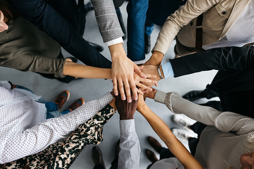 A high-angle shot of a group of male and female colleagues putting their hands together in an office. They are dressed in fashionable business clothes. Their faces are not visible, only their arms. Horizontal daylight indoor photo.