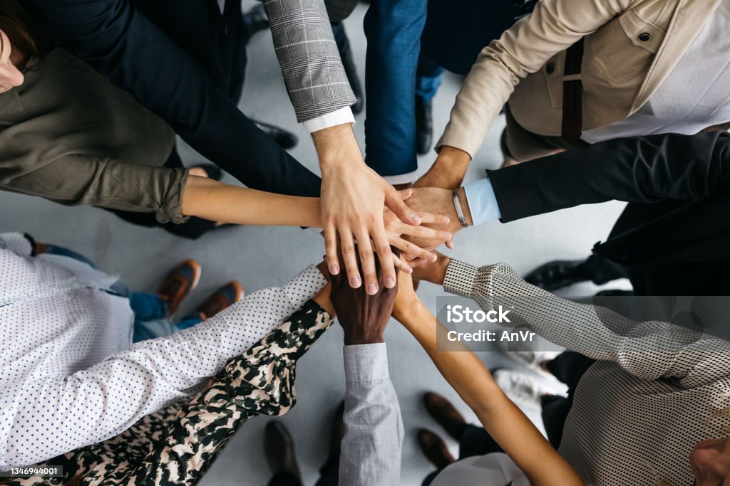 Close-up of co-workers stacking their hands together A high-angle shot of a group of male and female colleagues putting their hands together in an office. They are dressed in fashionable business clothes. Their faces are not visible, only their arms. Horizontal daylight indoor photo. Teamwork Stock Photo