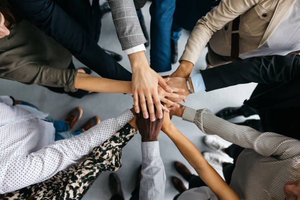 Close-up of co-workers stacking their hands together A high-angle shot of a group of male and female colleagues putting their hands together in an office. They are dressed in fashionable business clothes. Their faces are not visible, only their arms. Horizontal daylight indoor photo. togetherness stock pictures, royalty-free photos & images