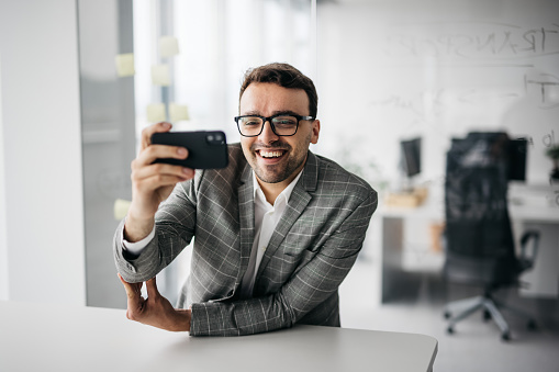 A young smartly-dressed businessman is having a videocall on his mobile phone. He is smiling at the camera and having fun. The office he works in is modern and bright. Horizontal daylight photo.