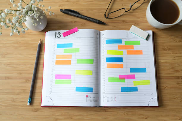 Open diary or appointment calendar with a lot of colorful sticky notes on a wooden desk with coffee pens and glasses, time management concept, copy space, selected focus, flat lay stock photo