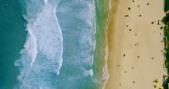 Aerial locked shot of waves breaking on the shore. Colourful beach umbrellas and people enjoying the summer.