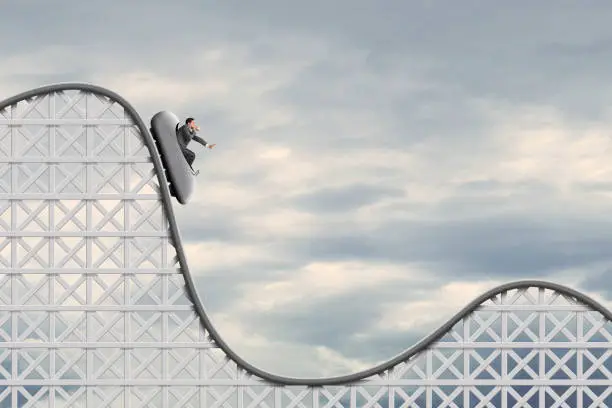 A businessman places a hand out in front of him and breathes into a paper bag as he sits in a rollercoaster car before as it begins a plunge.