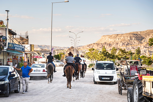 June 18, 2021, Turkey, Cappadocia - Picturesque landscape of Goreme. Riders on horses pass between cars on a city street against the backdrop of mountains
