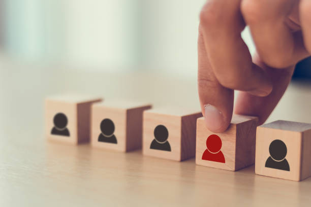 Human resource management, recruitment and target customer business concept. Man's hand puts wooden cube red person which standing on crowd with black person symbol. Bright background and copy space. gestión de recursos humanos, reclutamiento y concepto de negocio de cliente objetivo. la mano del hombre pone cubo de madera de la persona roja que de pie sobre la multitud con el símbolo de la persona negra. fondo brillante y espacio de copia.