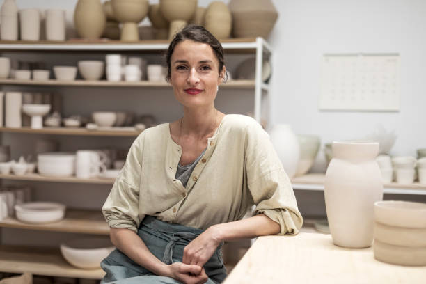 porträt einer schönen glücklichen handwerkerin, die schürze trägt und lächelt, während sie in ihrem kunstatelier oder bastelladen sitzt - keramiker stock-fotos und bilder