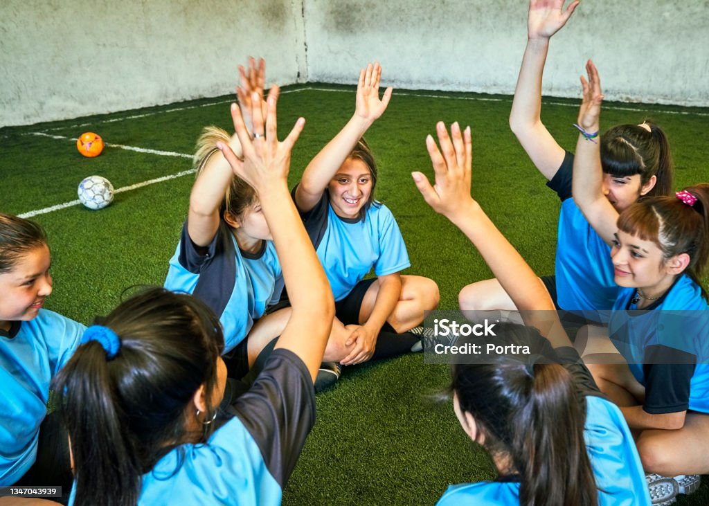 Female soccer team motivating each other before game Girls soccer team players in blue jerseys getting ready for game, they are sitting at sports court with their hands raised and smiling. Sports Training Stock Photo