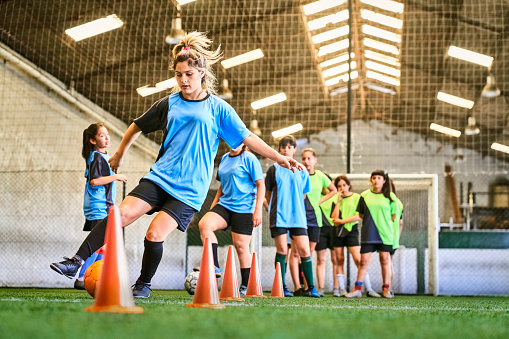 Group of girls on soccer training at indoor soccer court, doing dribbling drills. Teenage girl in blue jersey kicks soccer ball around orange cones.